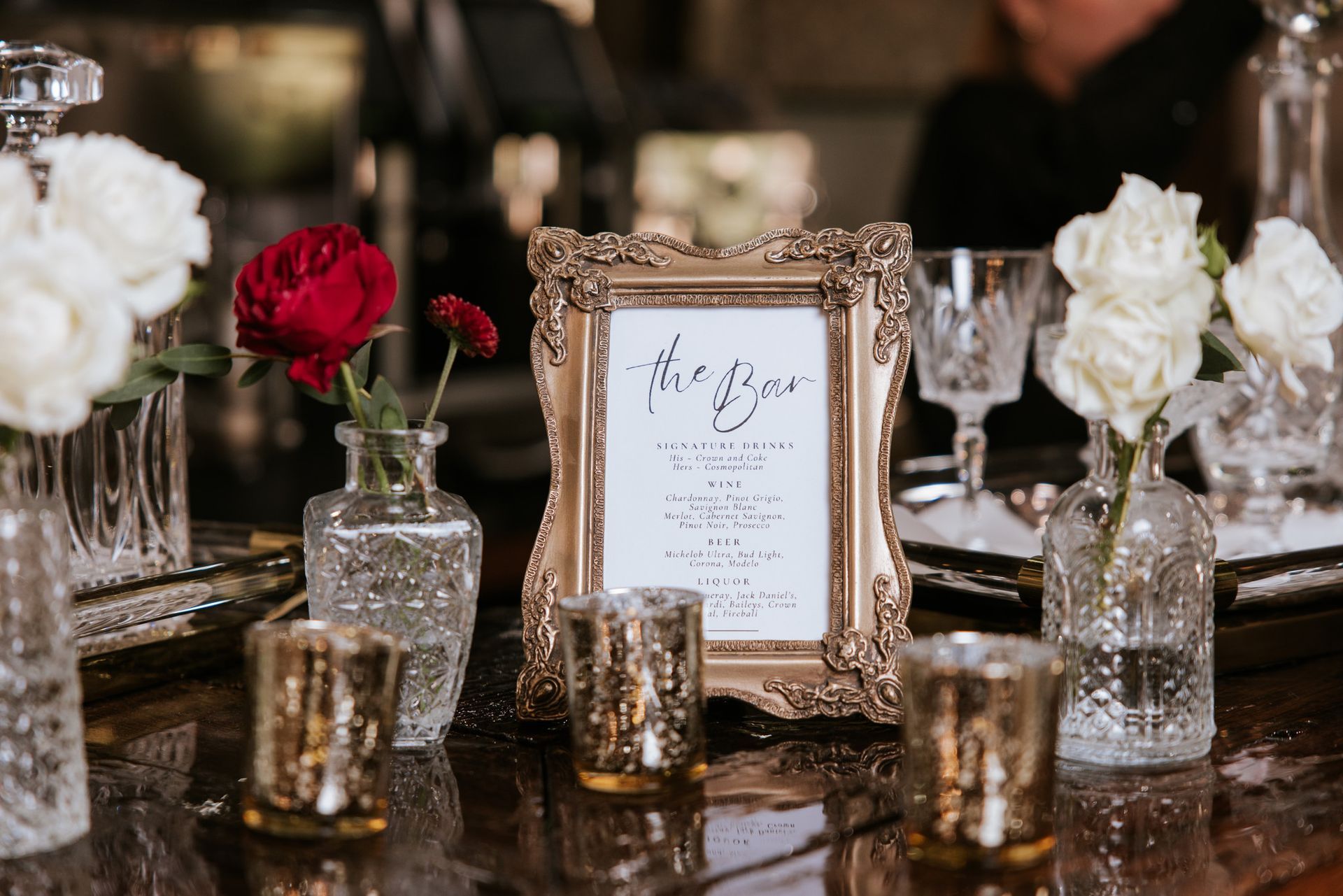 Elegant table centerpiece with white and red flowers, gold candle holders, and a framed menu card