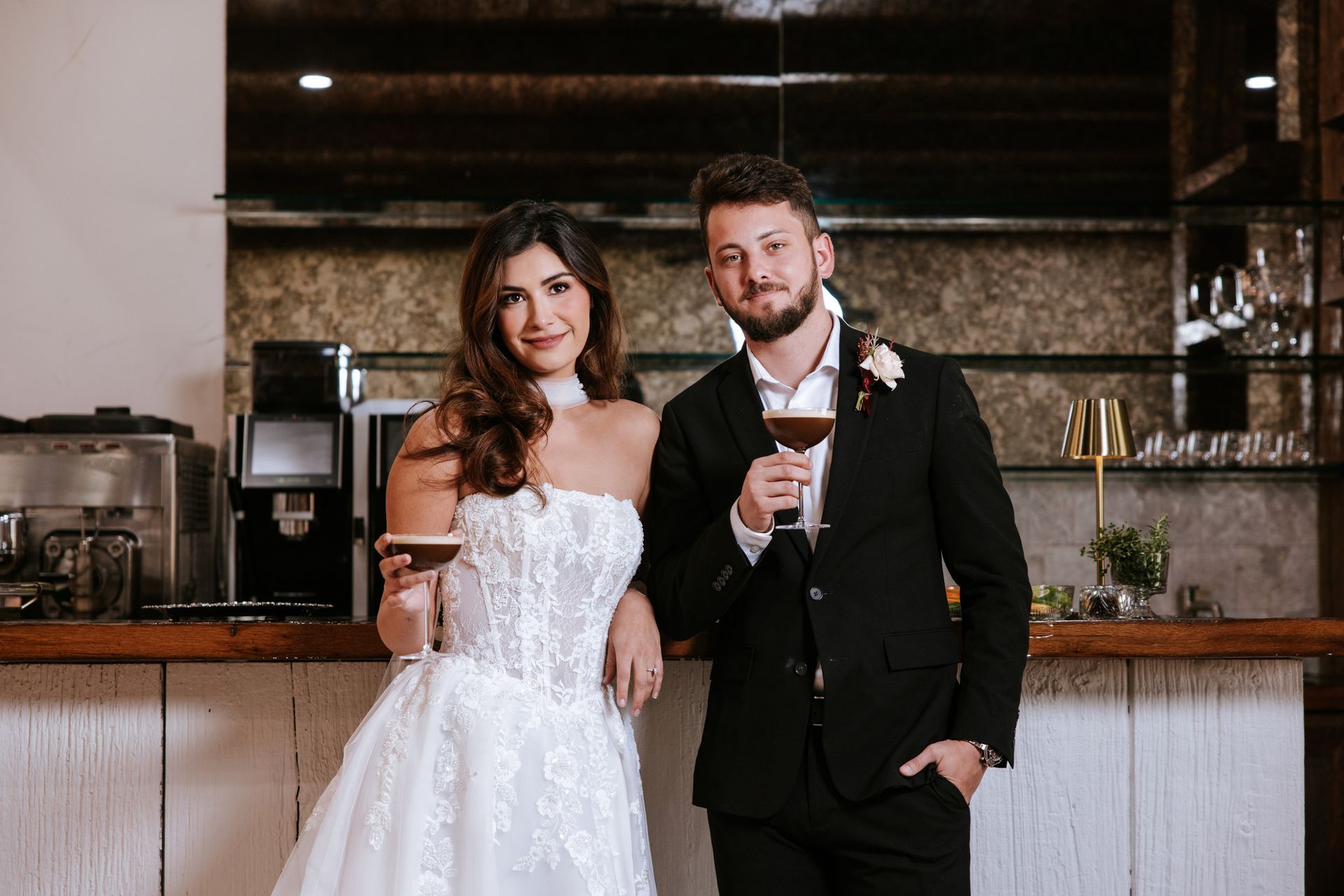 Bride in a white dress and groom in a black suit pose with drinks at a bar.