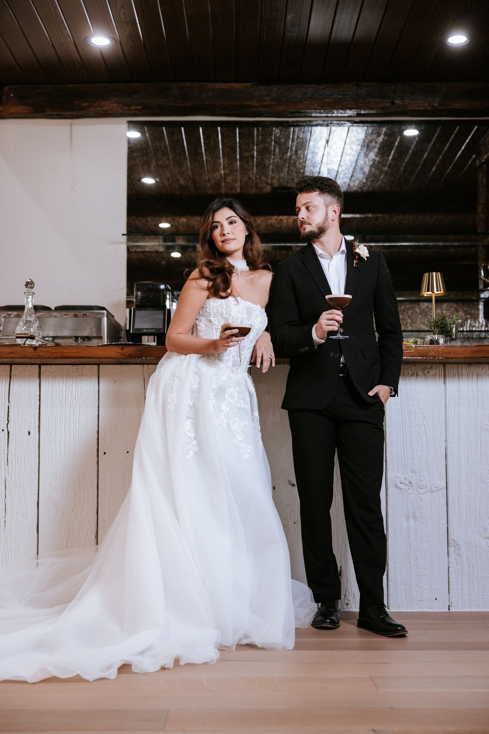 Bride and groom in formalwear standing at a bar, holding drinks, in a rustic indoor venue.