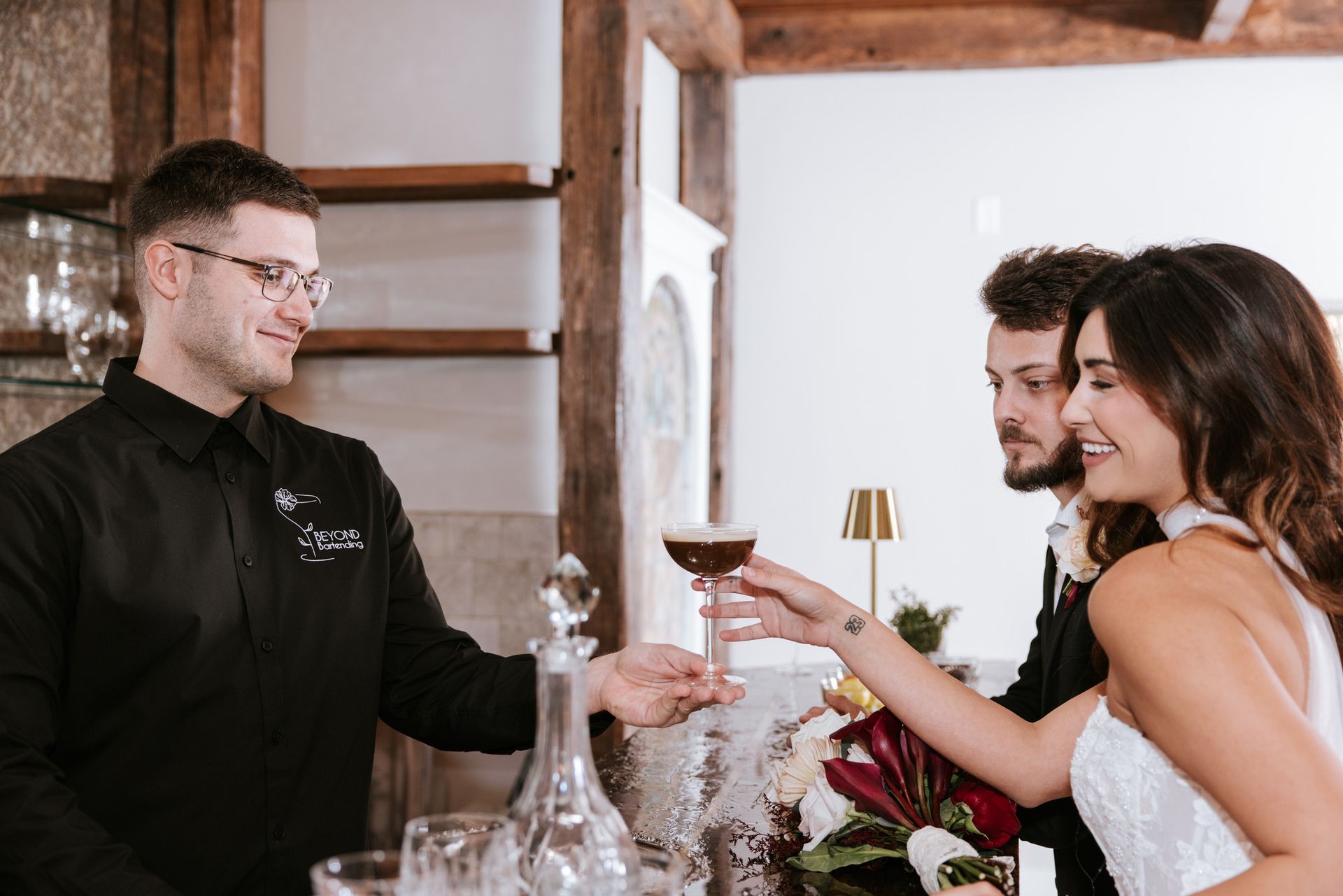 Bartender pours a drink for a smiling couple at a wedding reception bar.