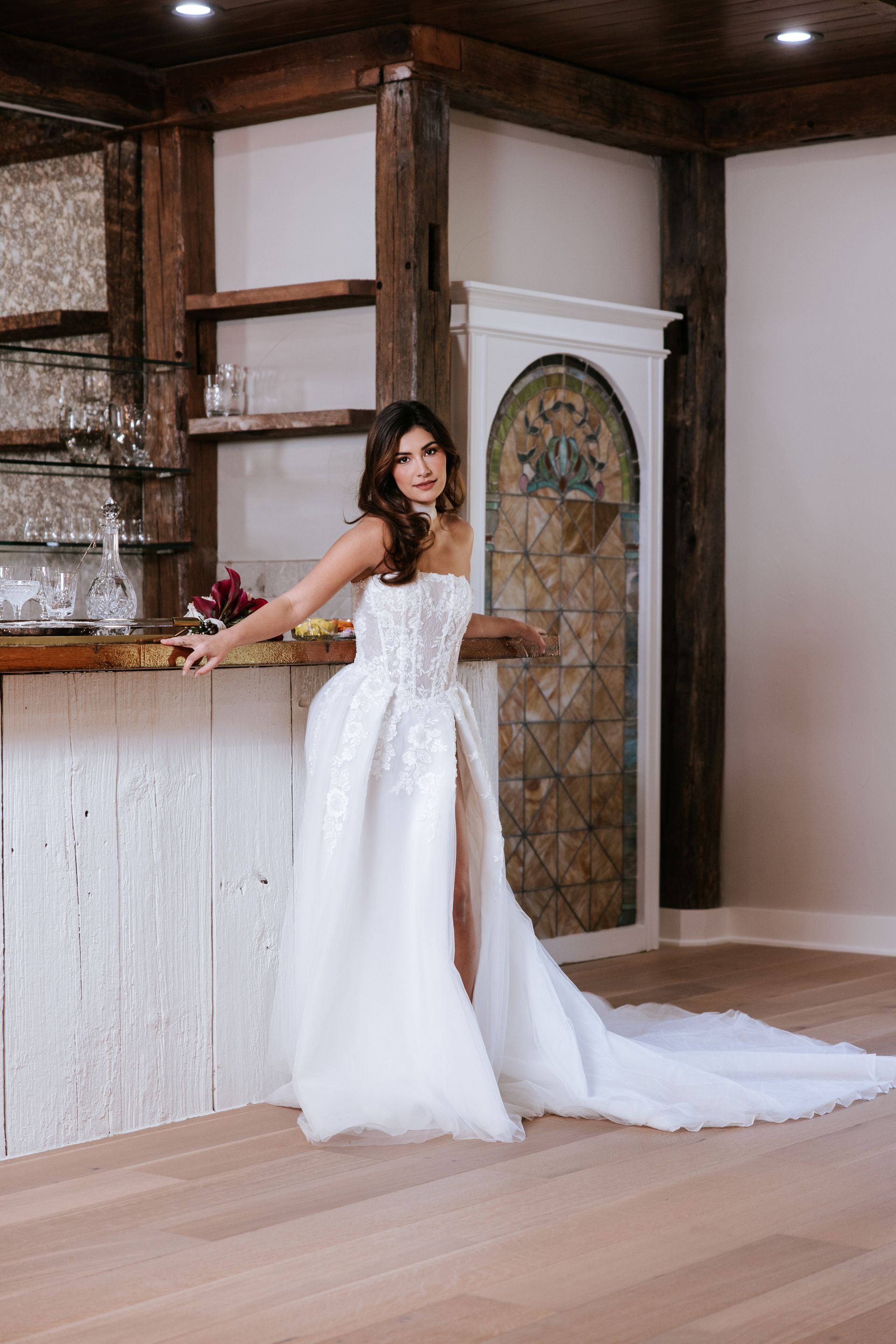 Woman in a white gown standing by a rustic bar in a wood-paneled room.