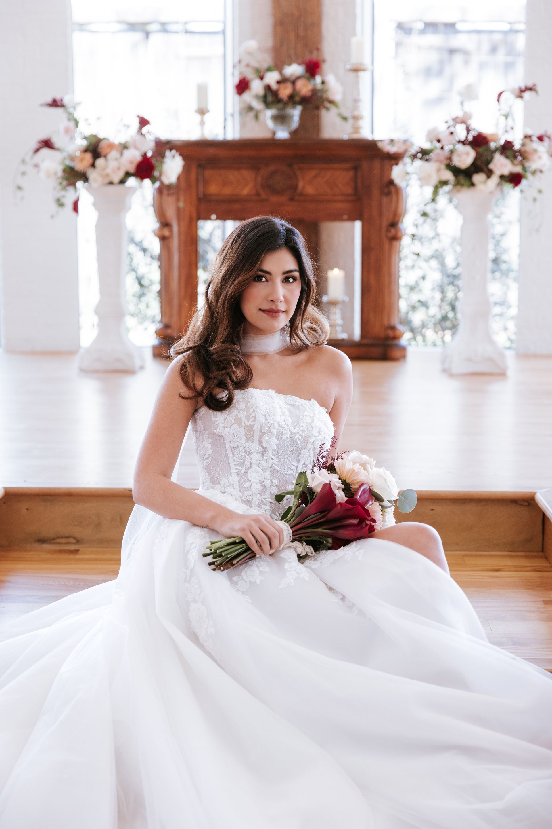 Bride in a white gown seated in a bright wedding hall, holding a bouquet with a floral altar behind her