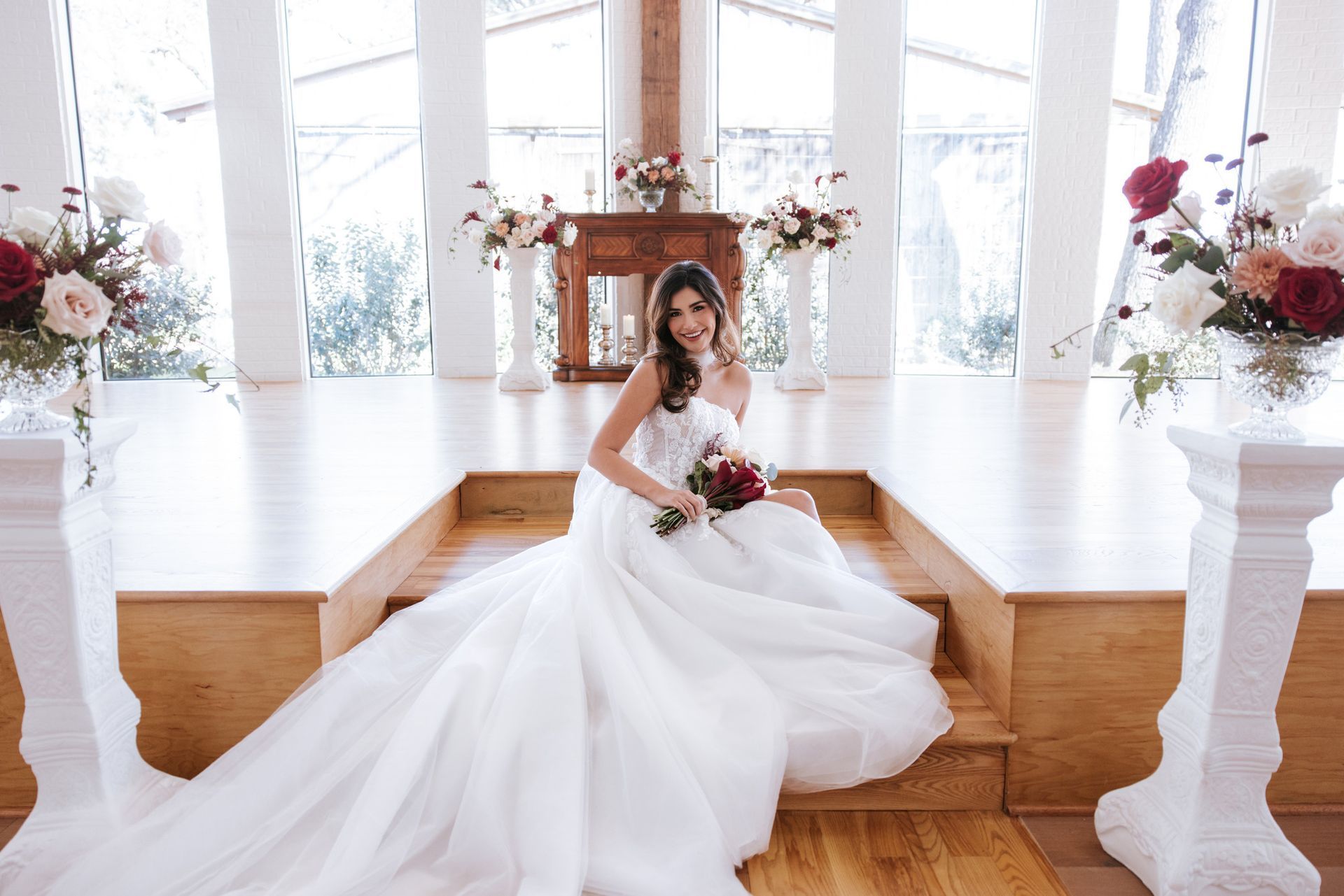 Bride in white gown seated on a wooden stage holding a bouquet in a bright wedding hall