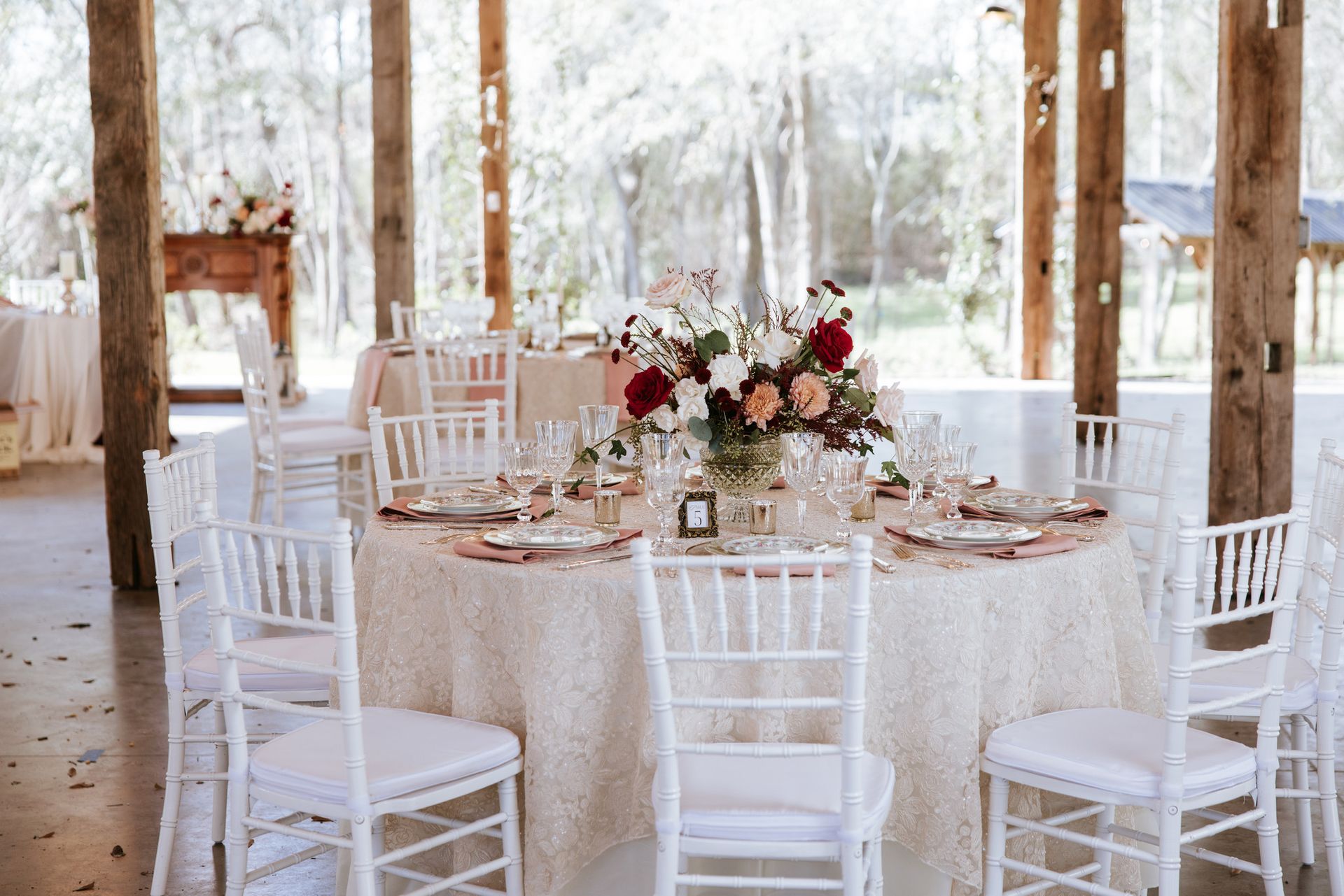 Round wedding table with white chairs, lace tablecloth, floral centerpiece, and rustic wooden pavilion backdrop