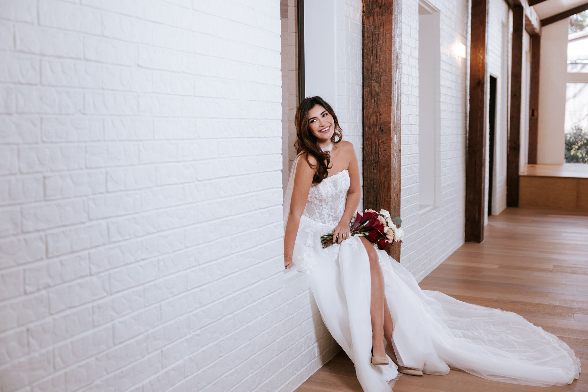 Bride in a white gown sitting by a white brick wall in a hallway, holding a bouquet and smiling.