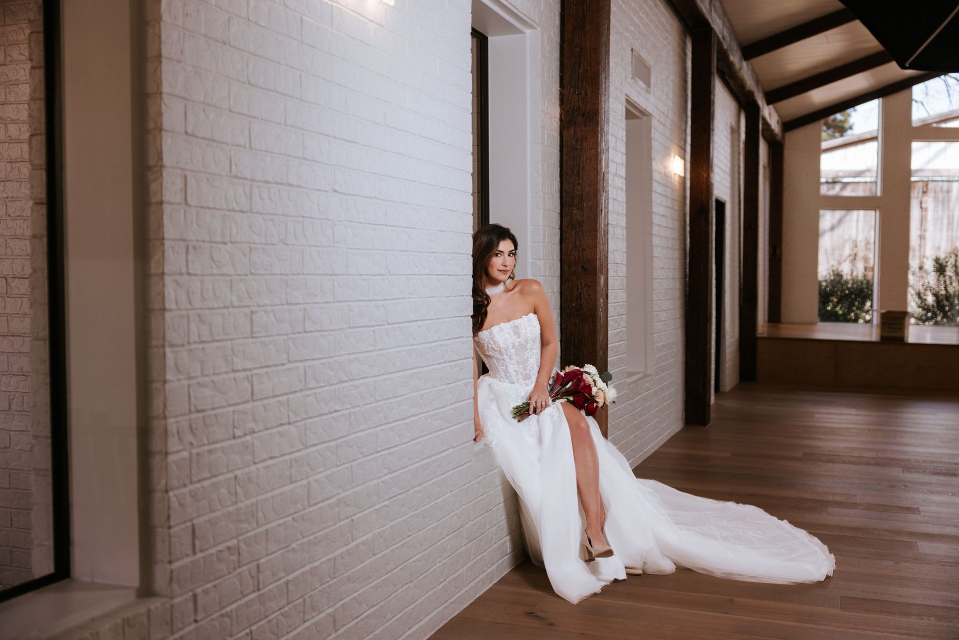 Bride in white gown leaning against a hallway wall, holding a bouquet in a bright corridor.