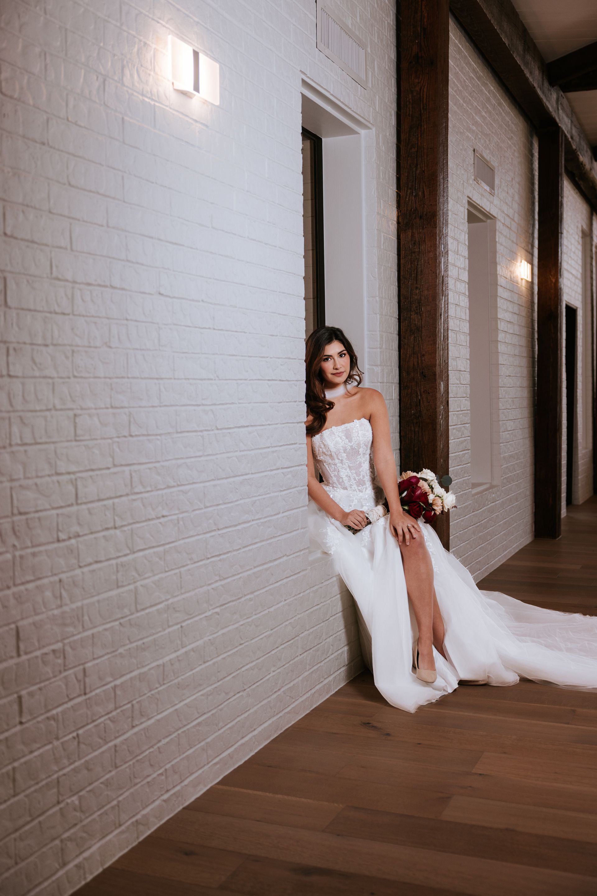 Bride in a white gown leaning against a white brick wall in a dim hallway, holding a bouquet.