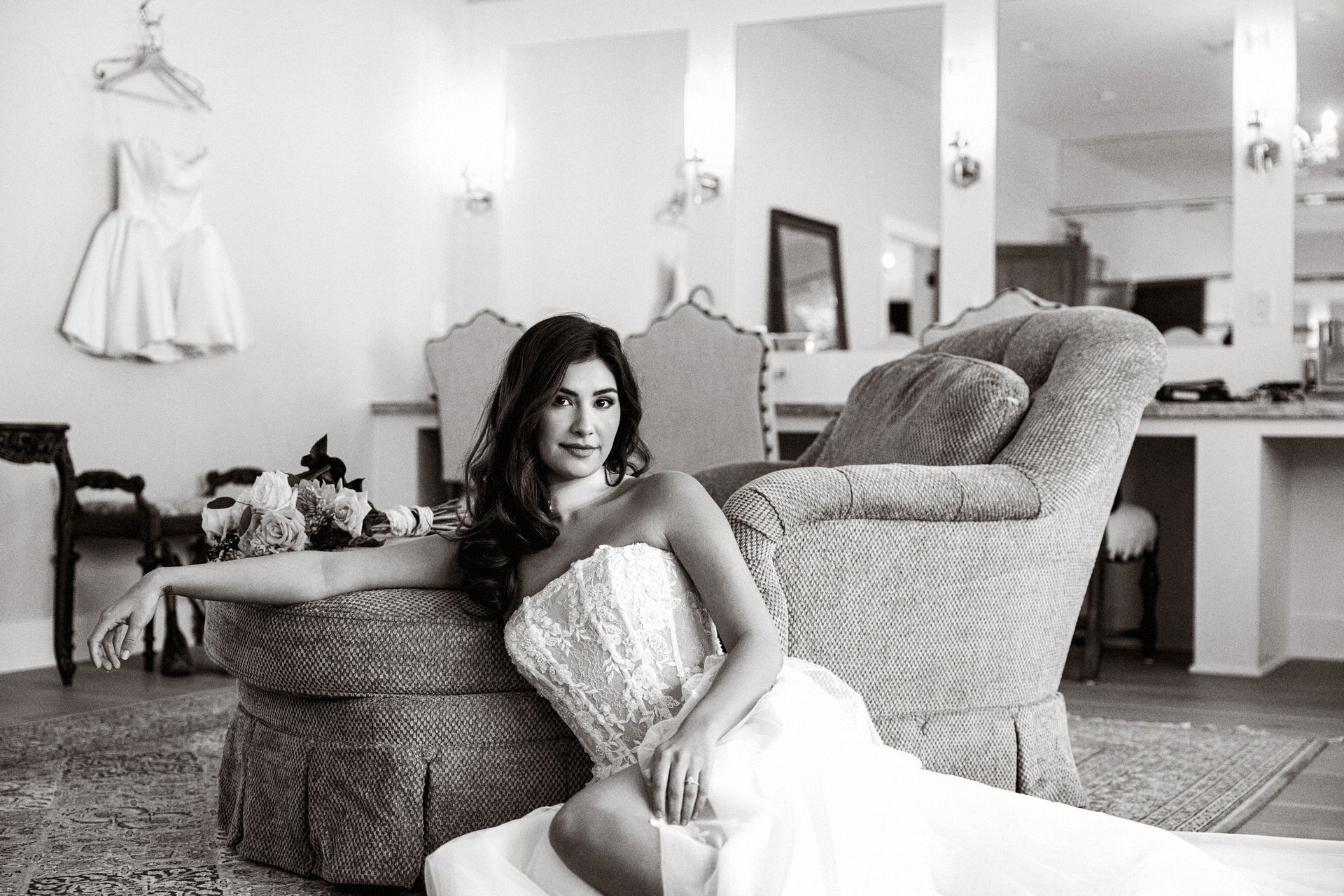 Woman in a white gown reclining on a wicker chair in a bright dressing room, with a large mirror and vanity behind her