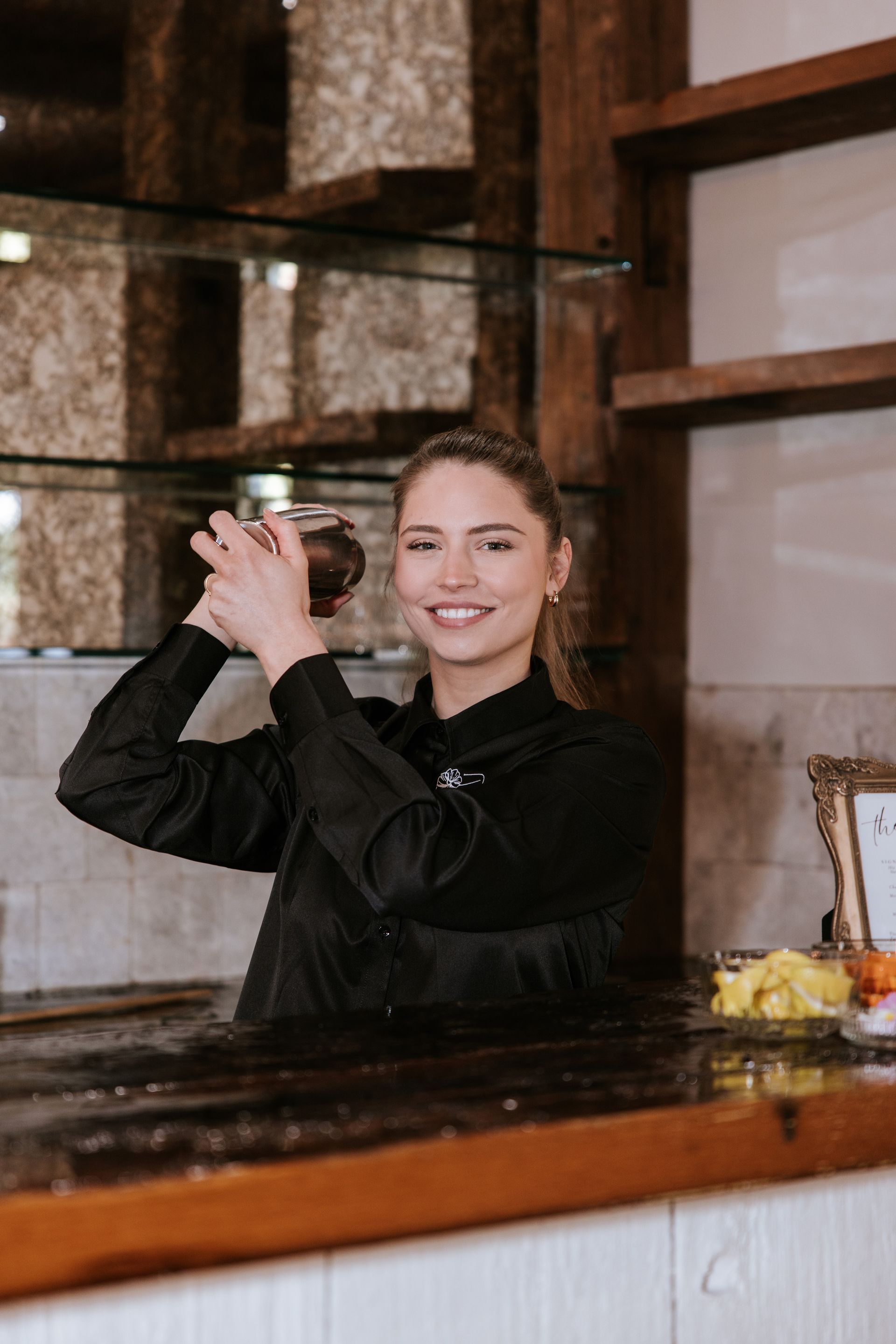 Smiling bartender in a black top shaking a cocktail behind a wooden counter