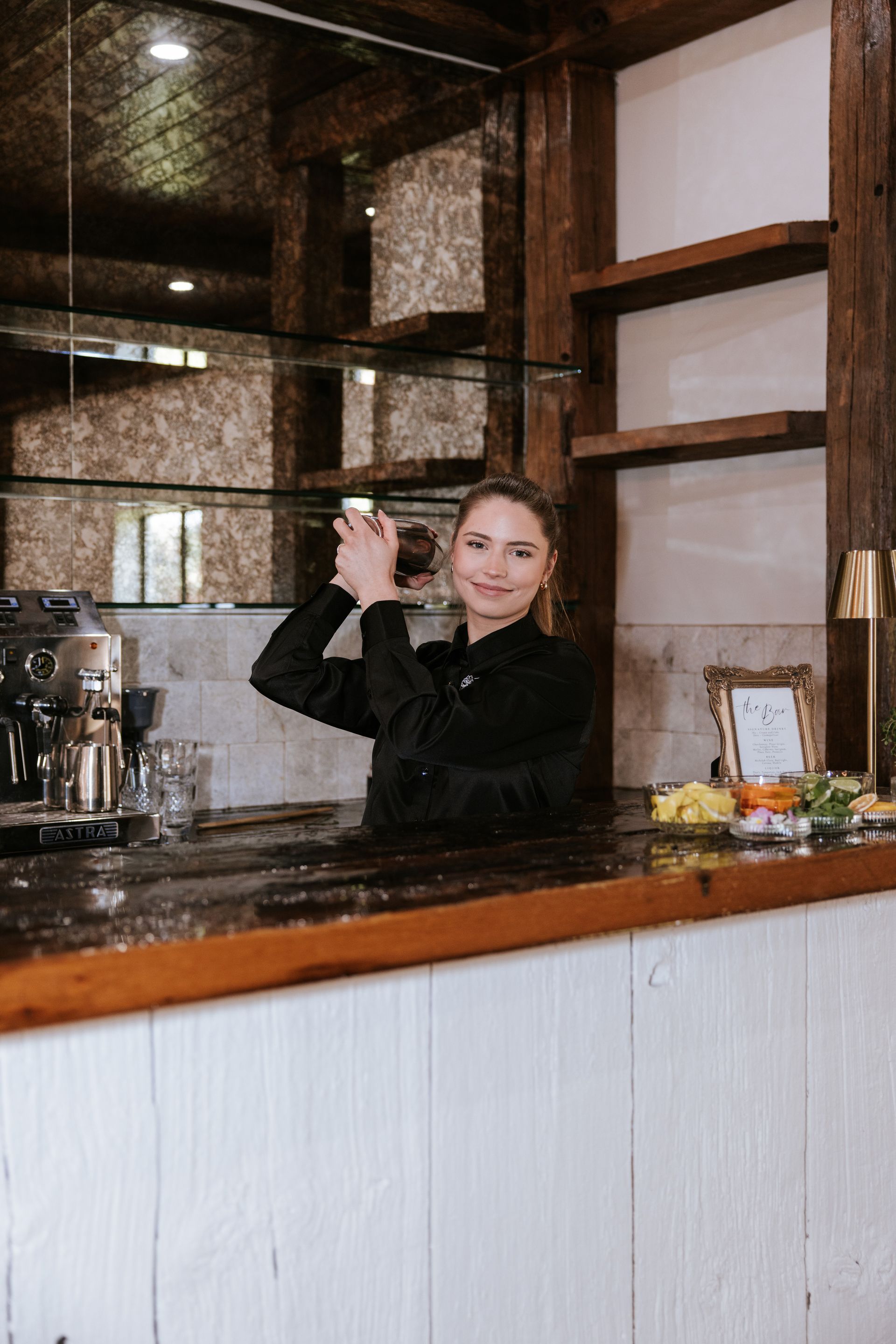 Woman behind a cafe counter smiling and waving, with coffee equipment and shelves in the background