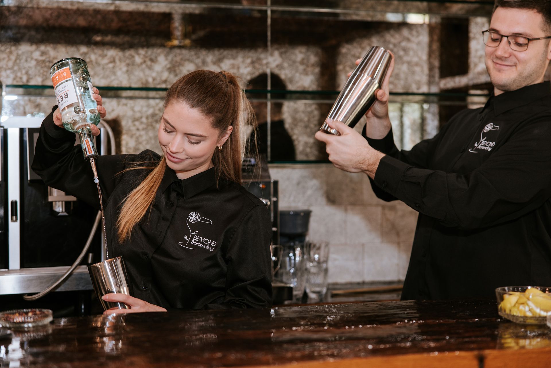Two bartenders pouring drinks behind a bar, one shaking a cocktail shaker and smiling.