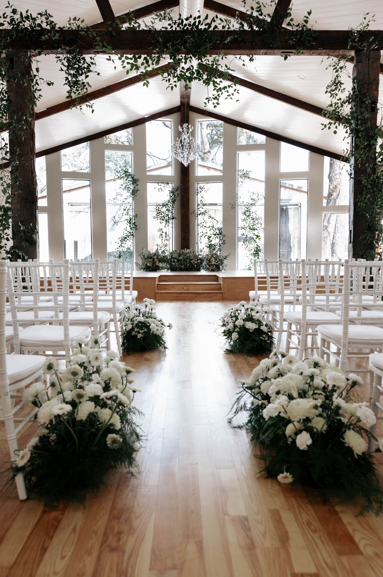 Wedding ceremony setup. White chairs line a wooden aisle adorned with white flower arrangements. Venue with large windows.