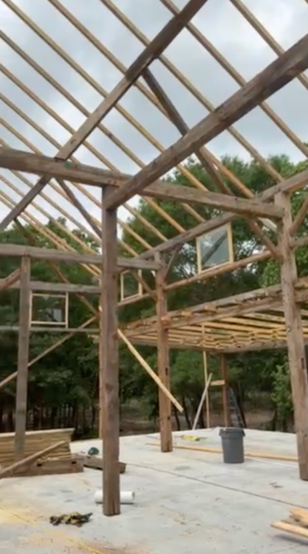 Wooden frame of a building under construction, with visible rafters and windows. Set outdoors, cloudy sky.