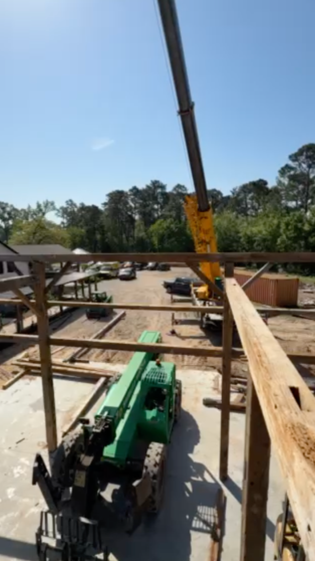 Construction site with a forklift lifting a wooden beam; blue sky and trees in the background.