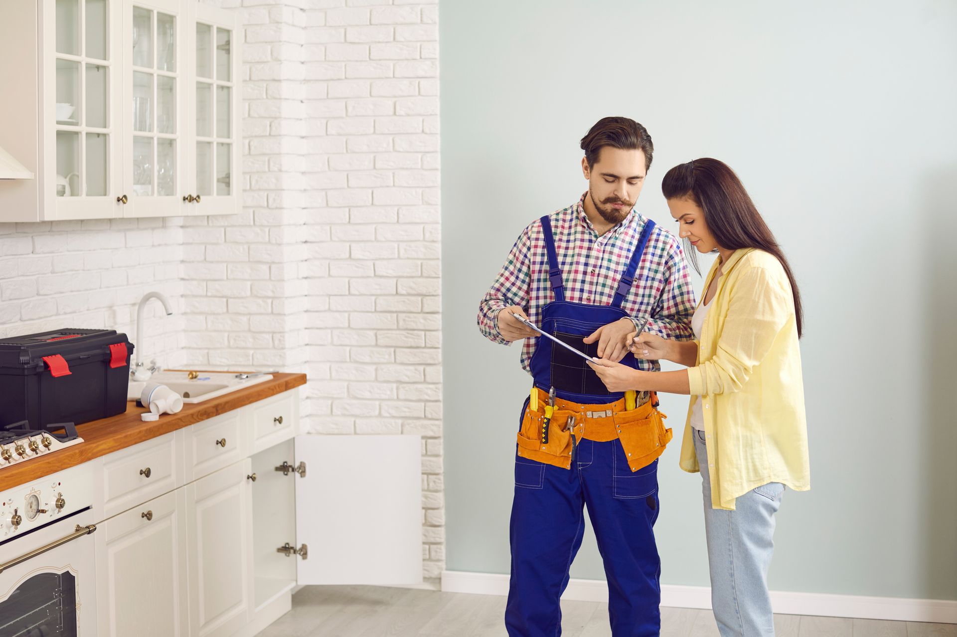 A plumber with tools and a clipboard explaining something to a woman in a kitchen.