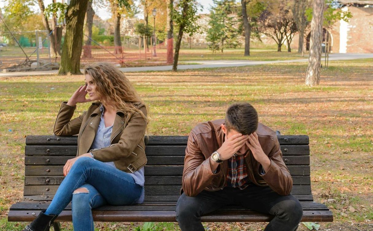 A man and a woman sitting on a bench in a park looking tense