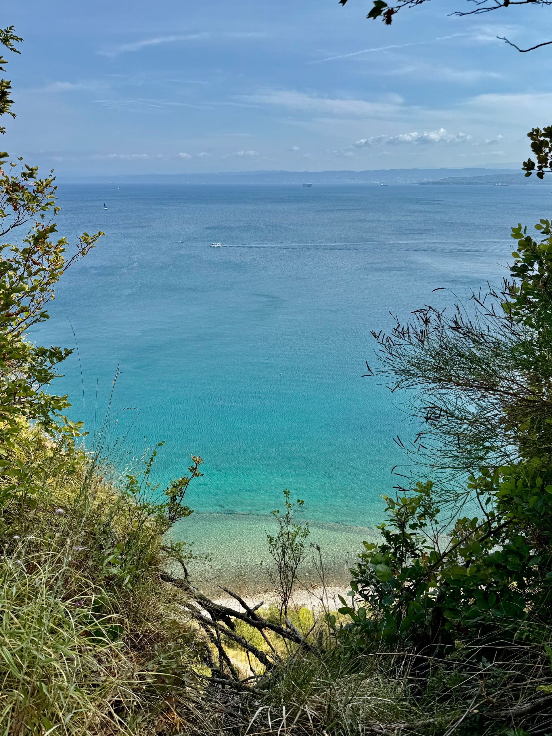 Ocean view from a leafy hillside, with turquoise water, distant coastline, and a sandy shore below. Bele Skale Beach, Slovenia