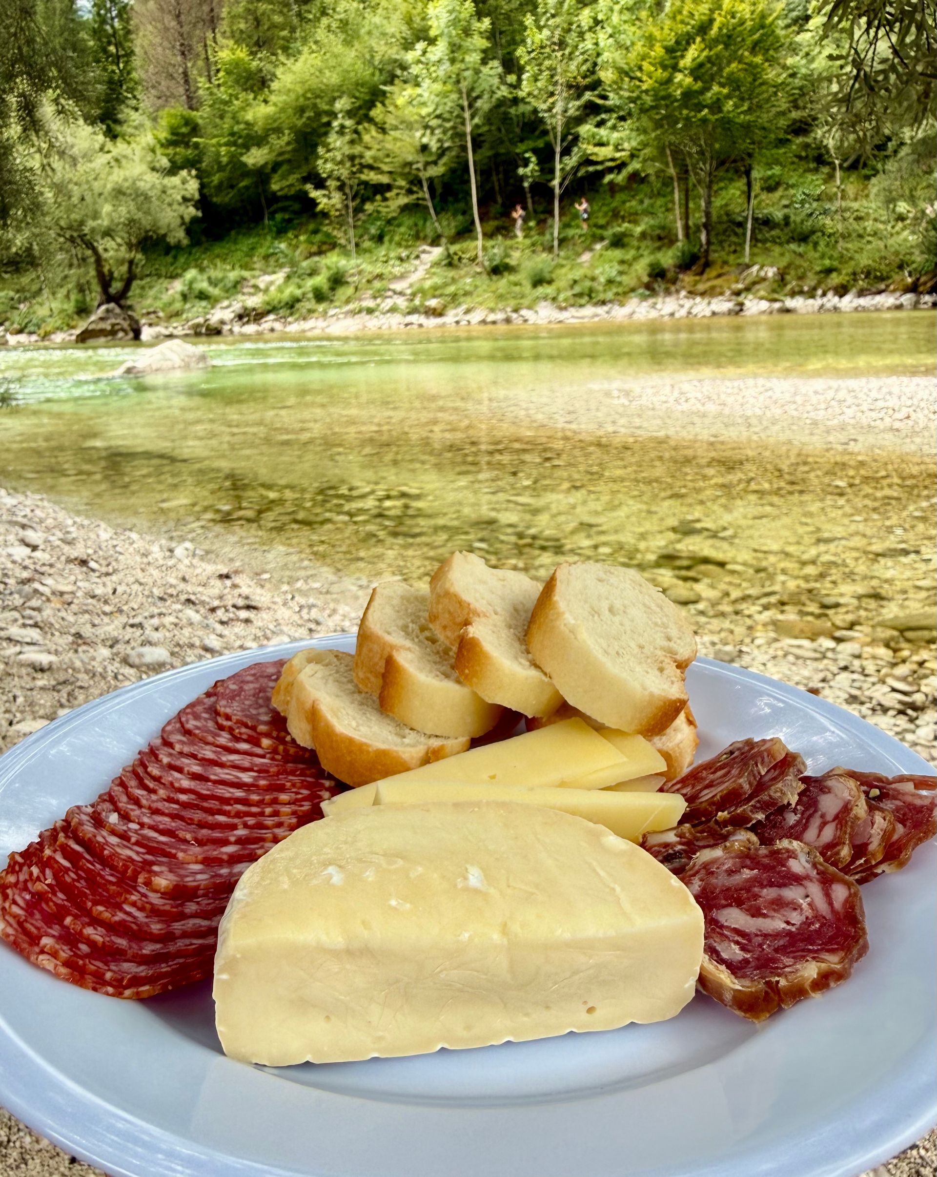 Picnic along the Soca River, Slovenia. Plate of assorted cheeses and sliced cured meats by a shallow river in a wooded outdoor setting
