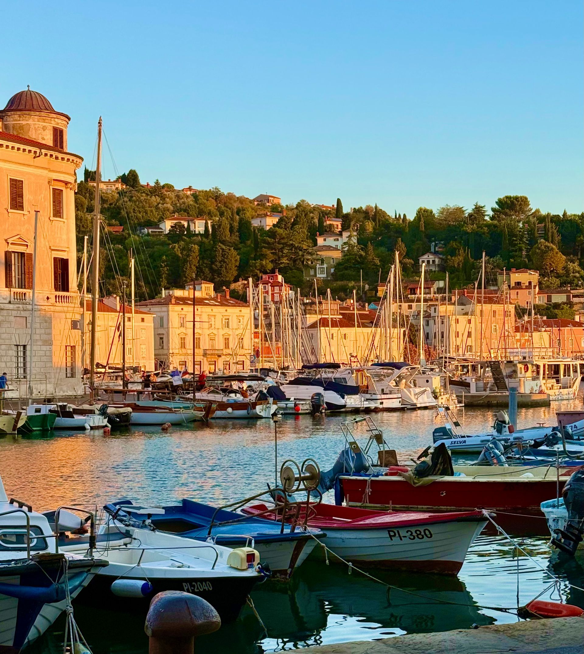 Sunlit harbor in Piran, Slovenia with moored boats, waterfront buildings, and a hillside town at Sunset