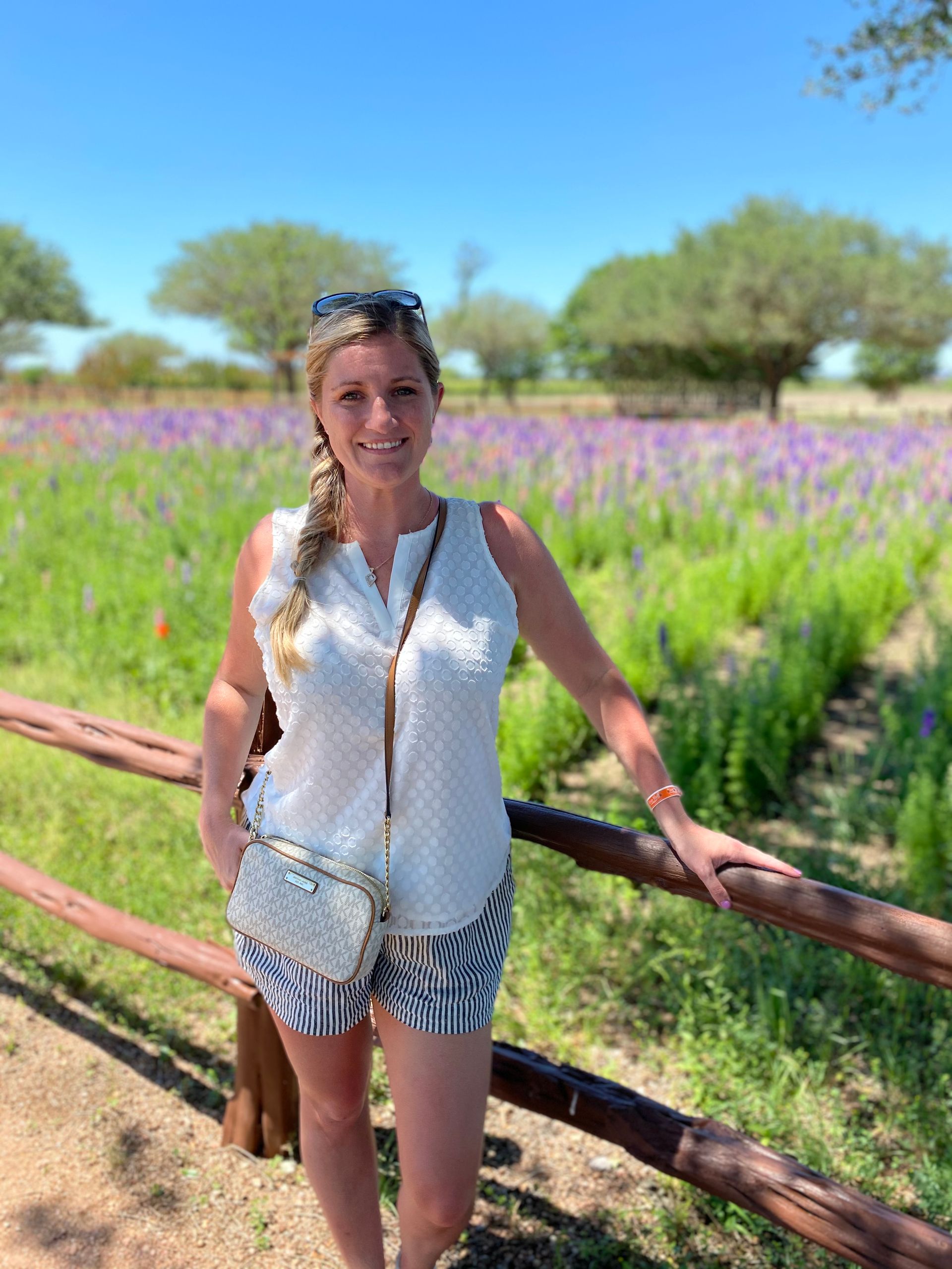 Woman in white top and striped shorts standing by a wooden fence in a purple flower field at wildseed farms in Fredericksburg Texas Wine Country