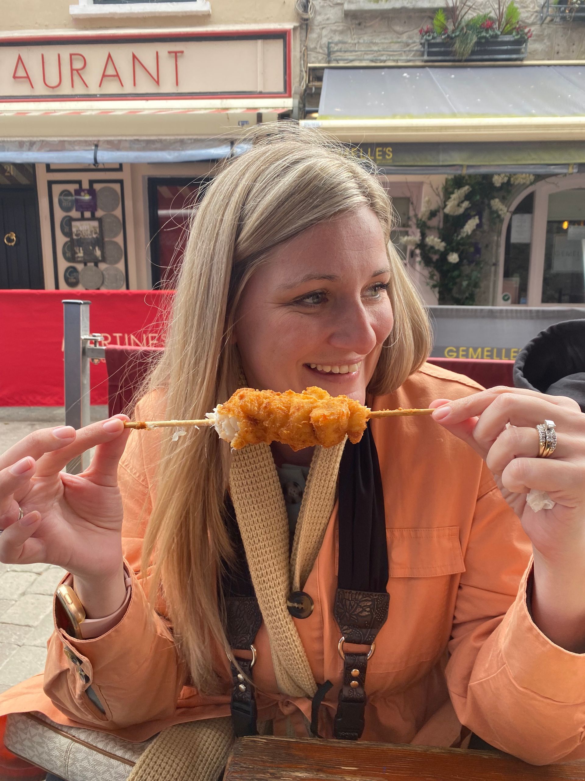 Woman in an orange jacket smiling while holding a skewer of grilled food at an outdoor Galway restaurant table