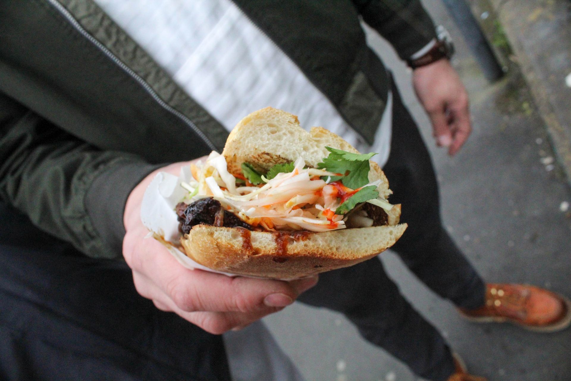 Hand holding an open sandwich with greens and sauce over a sidewalk at the Galway market