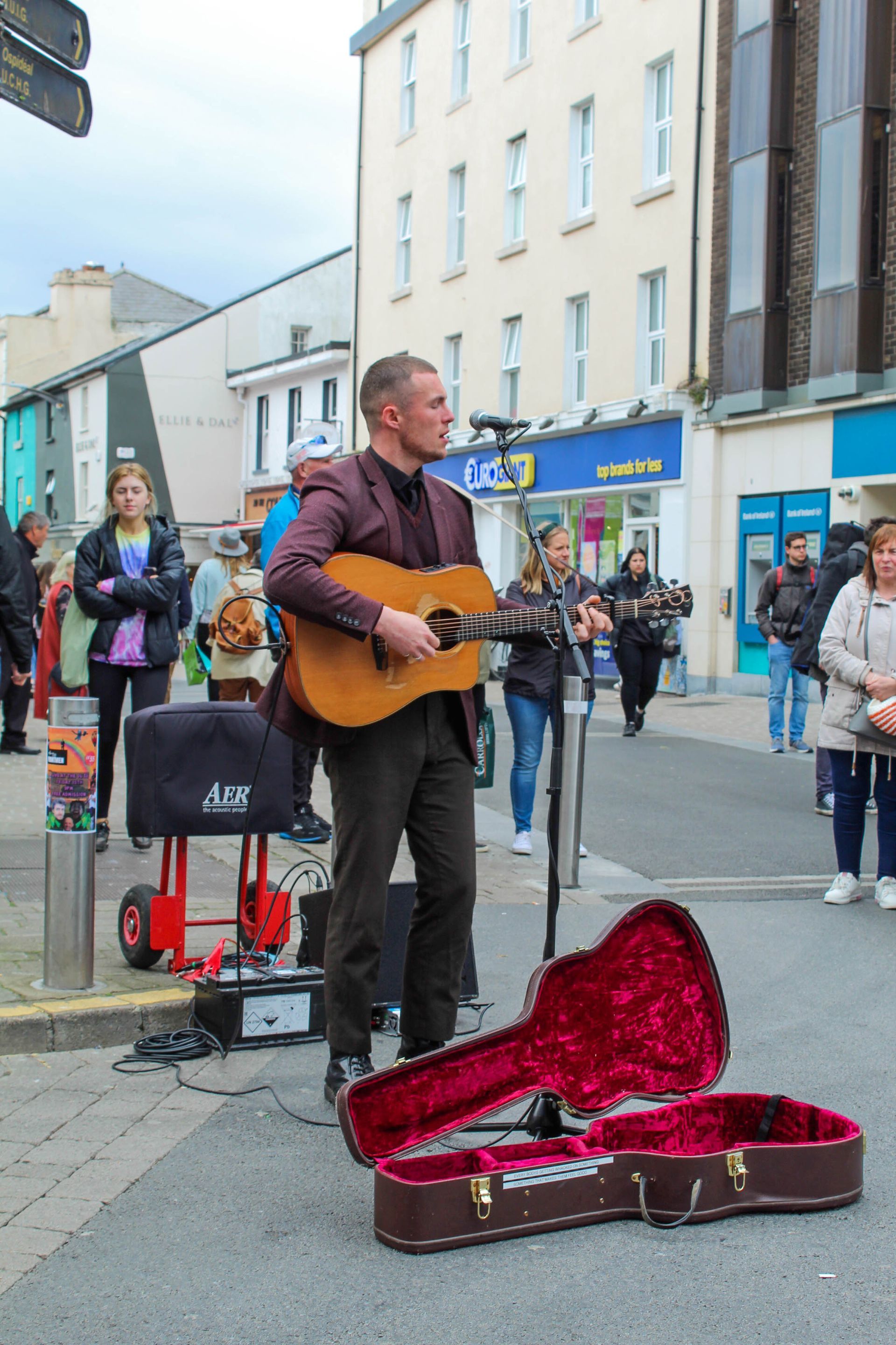 Street musician playing guitar on a Galway sidewalk, with an open red guitar case in front of him.