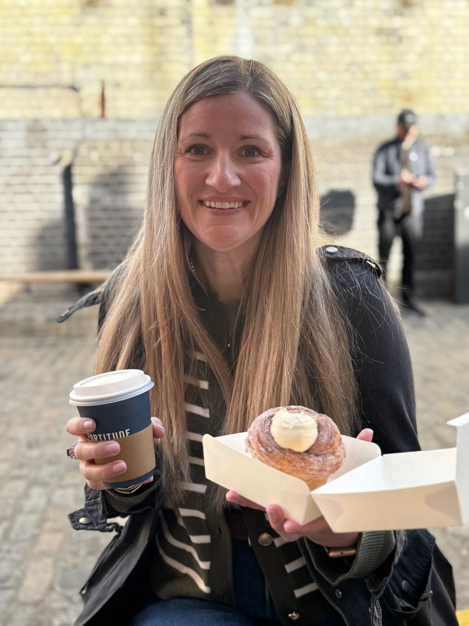 Smiling woman holding a coffee and a pastry outdoors in a sunlit plaza