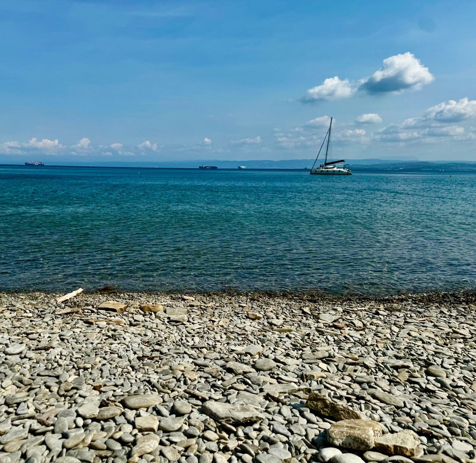 Rocky shoreline with turquoise sea and a sailboat under a blue sky at Bele Skale beach in Slovenia