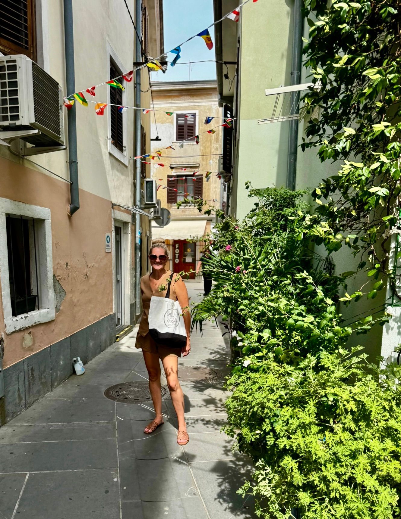 Woman walking down a narrow, colorful alley in Izola, Slovenia with greenery and hanging flags, carrying a Road and Table tote bag