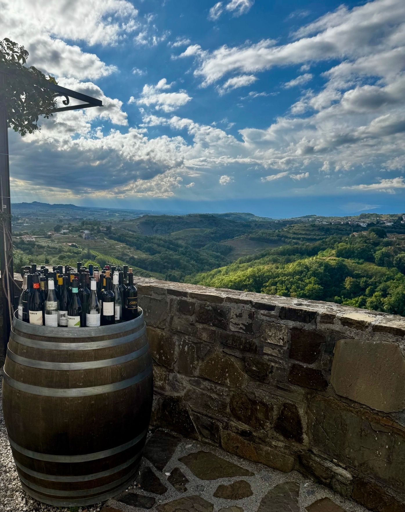Wine bottles in a barrel beside a stone wall overlooking a green valley and cloudy sky at Vinoteka Brda in Slovenia