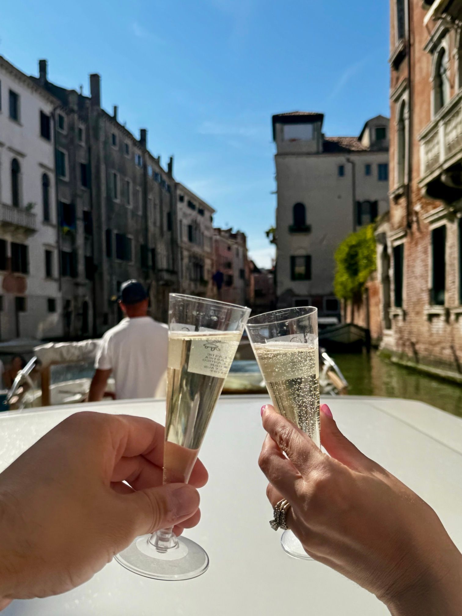 Hands clink champagne flutes on a sunny canal boat ride between Venetian buildings on the main canal in Venice. 