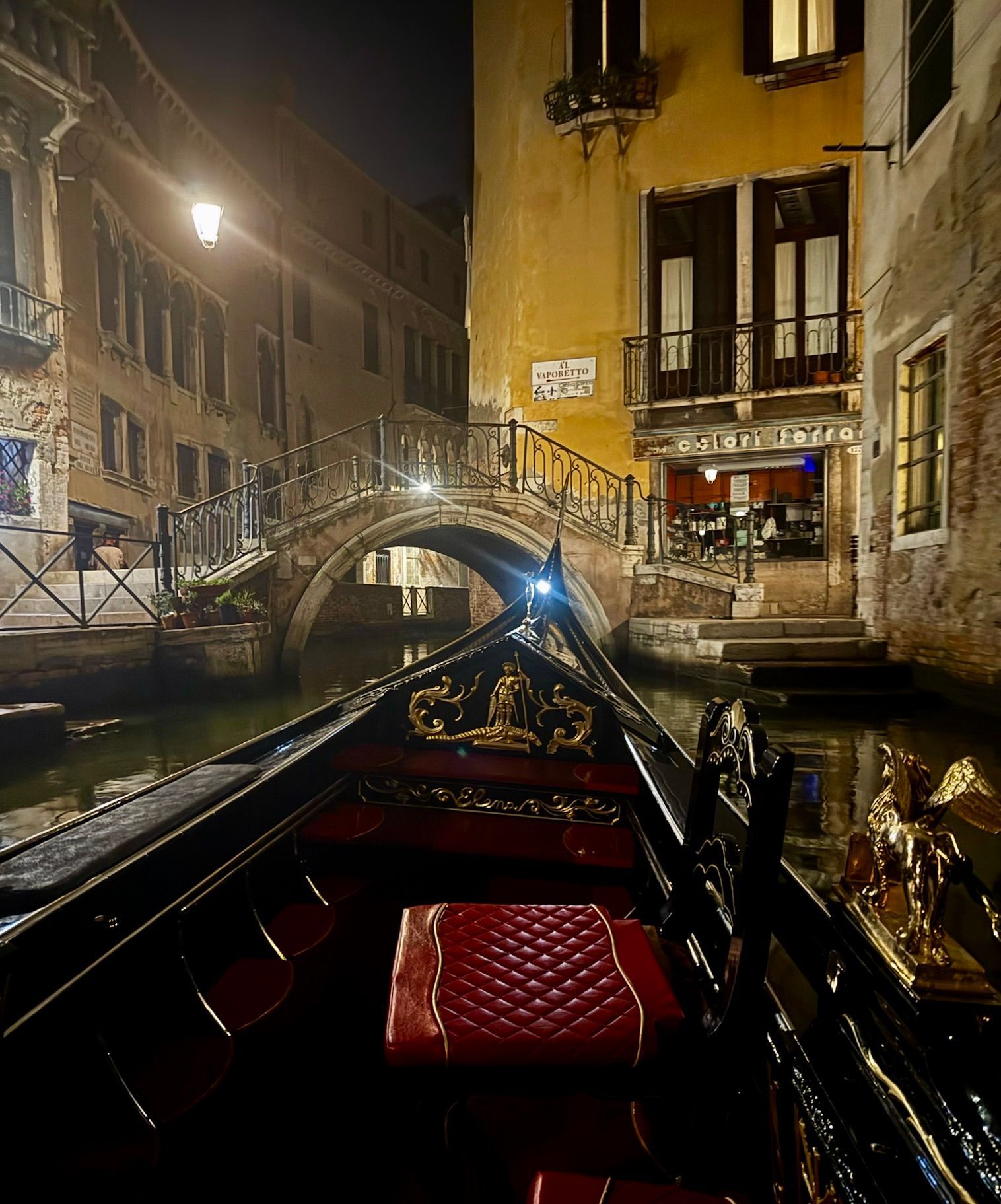 Night canal scene in Venice from a gondola, with a lit stone bridge and buildings reflected on dark water.