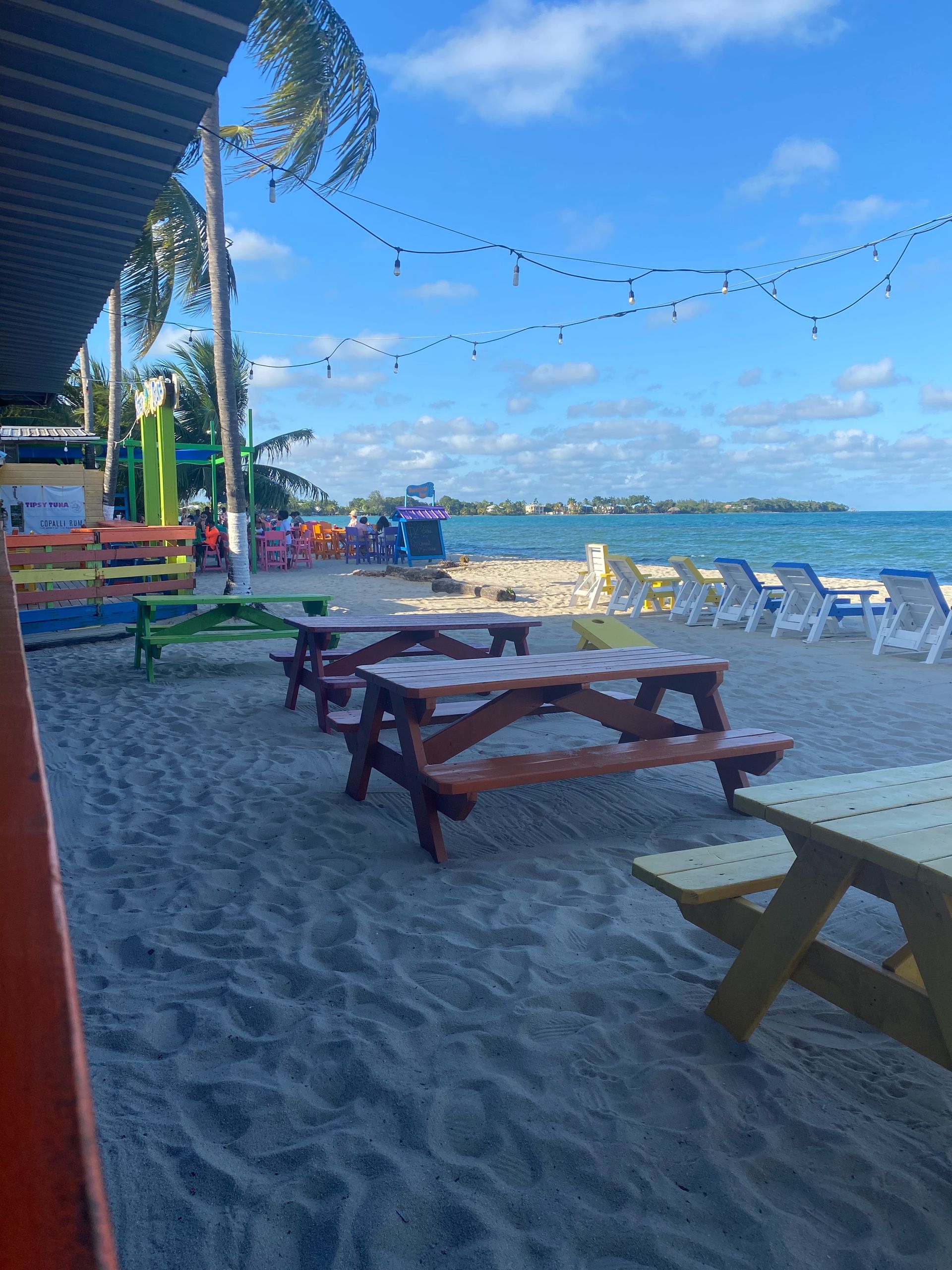 Beachside café seating on sand with picnic tables, string lights, and ocean waves under a clear blue sky