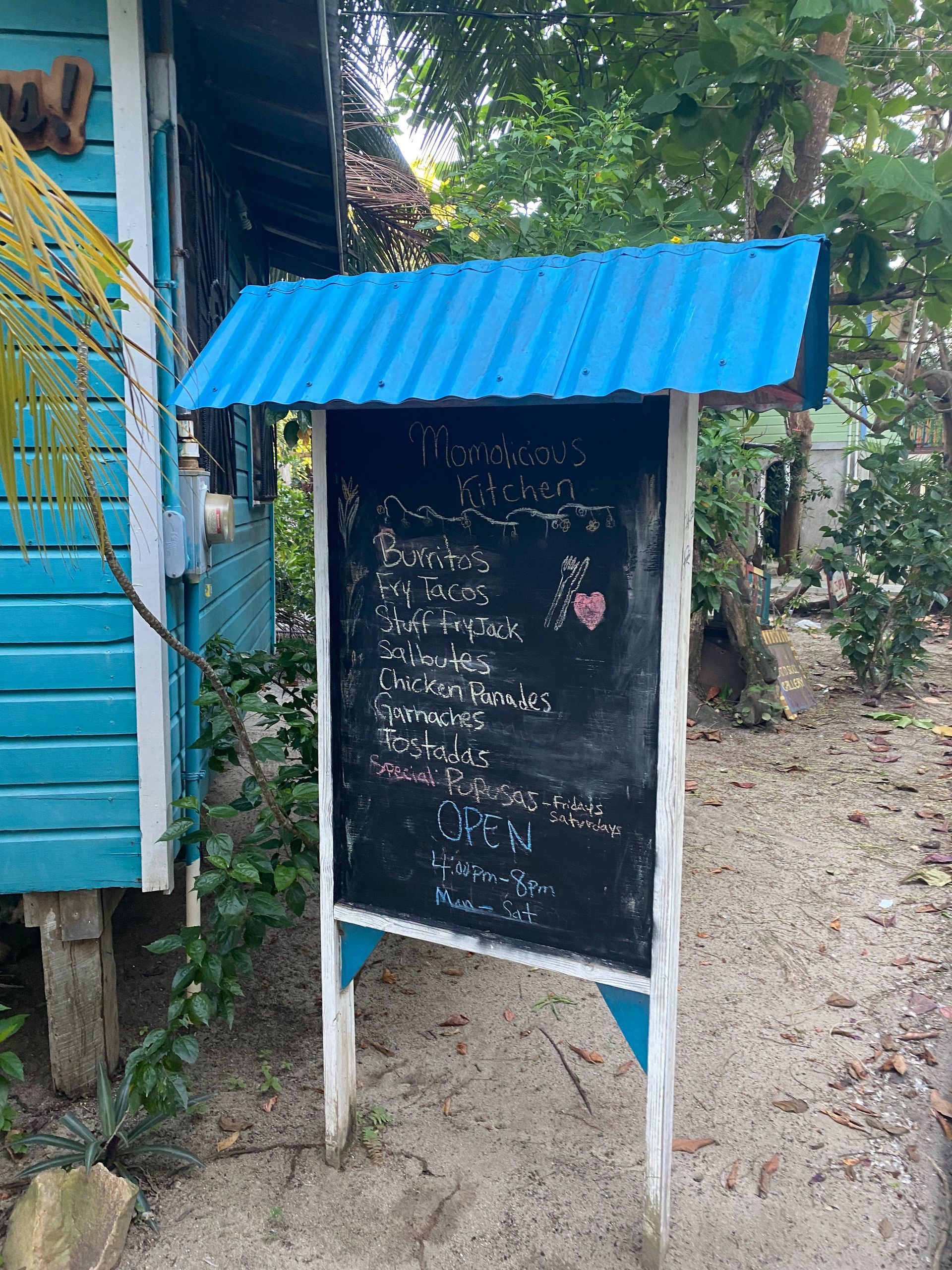 Black signboard included a chalk handwritten menu with blue roof stand outdoors beside a blue building and gravel path.