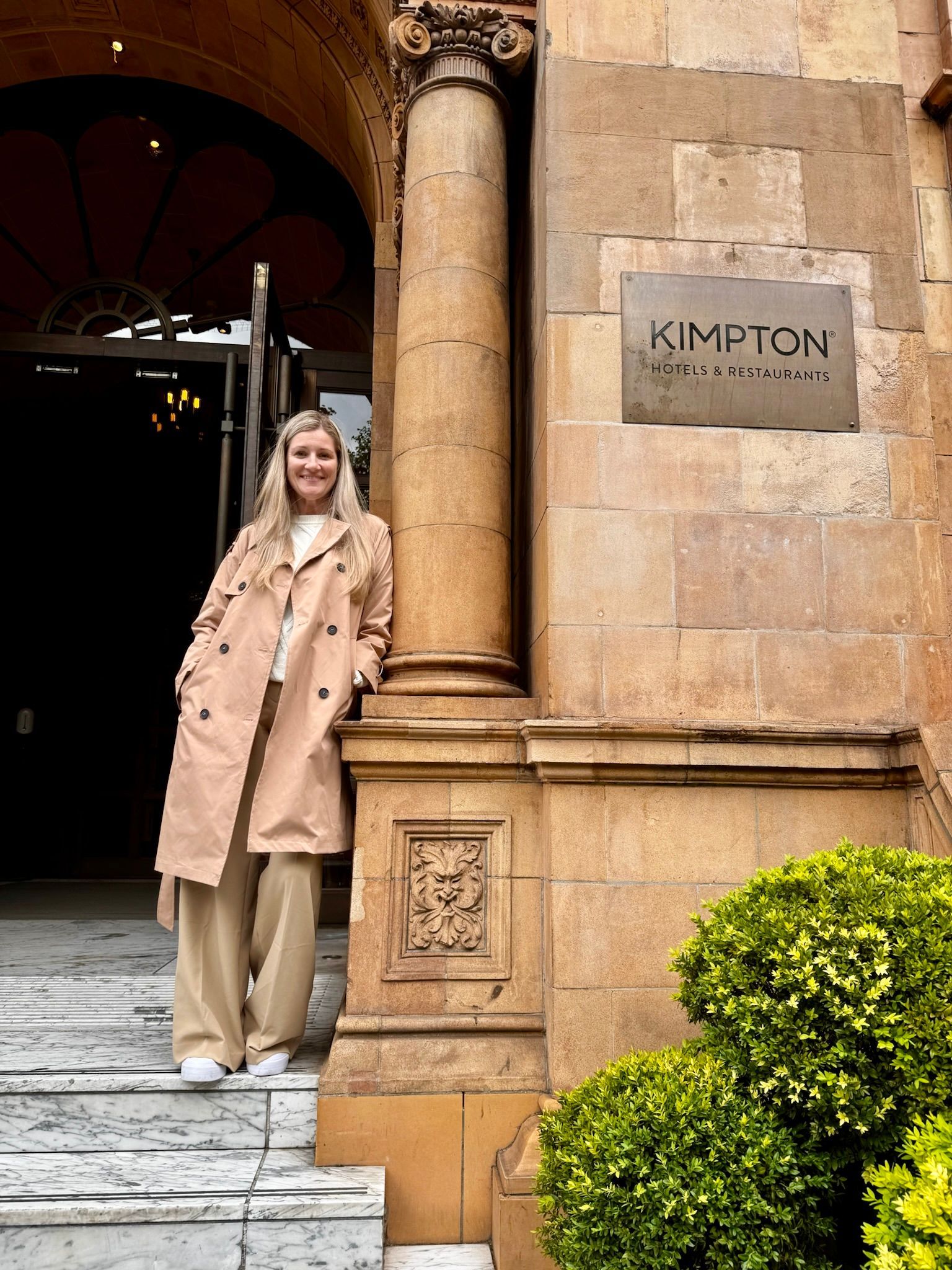 Woman in a beige coat standing by a Kimpton hotel entrance with stone columns and greenery