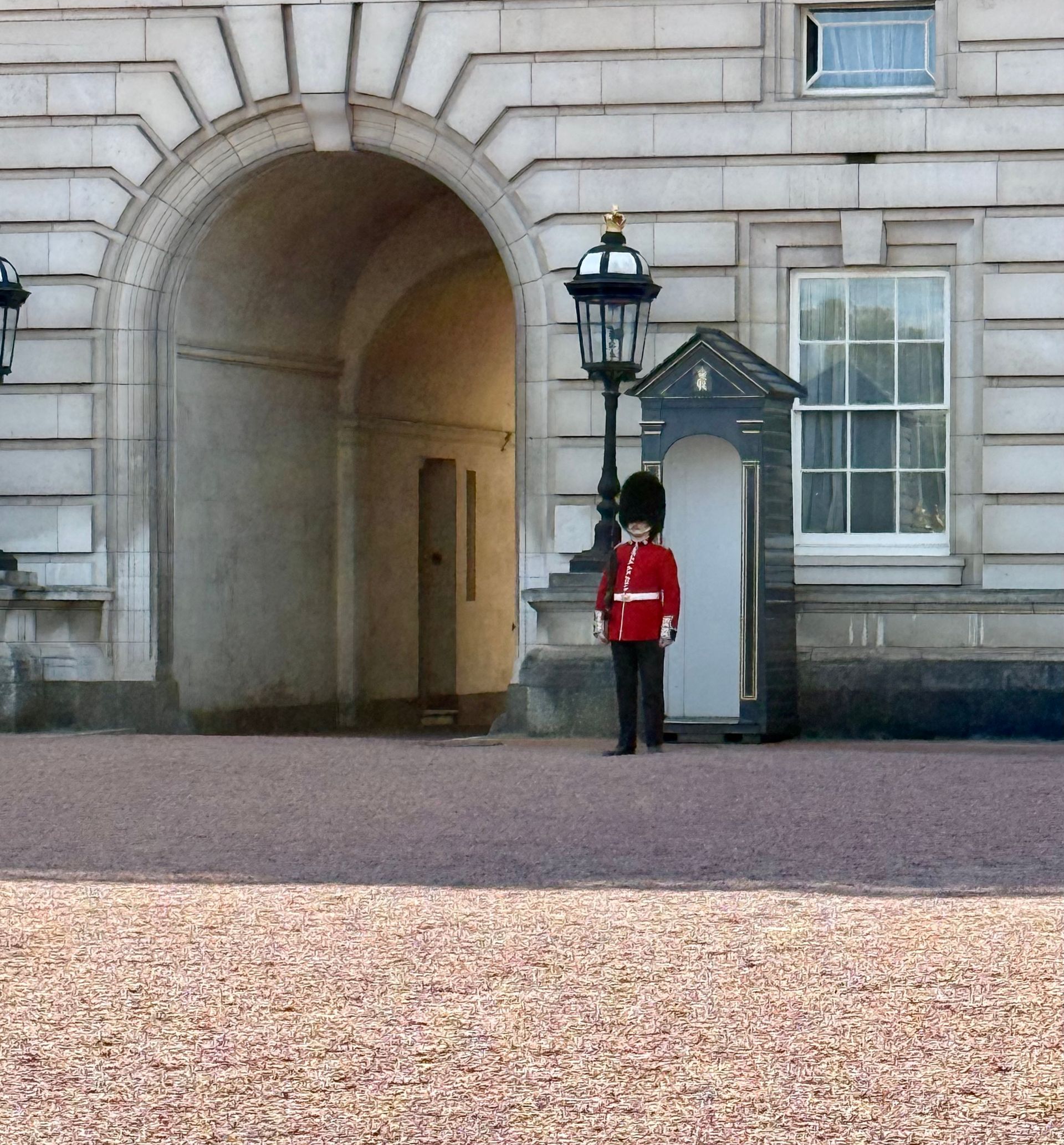 Guard in red uniform stands by a white sentry box beside a stone archway in front of Buckingham Palace
