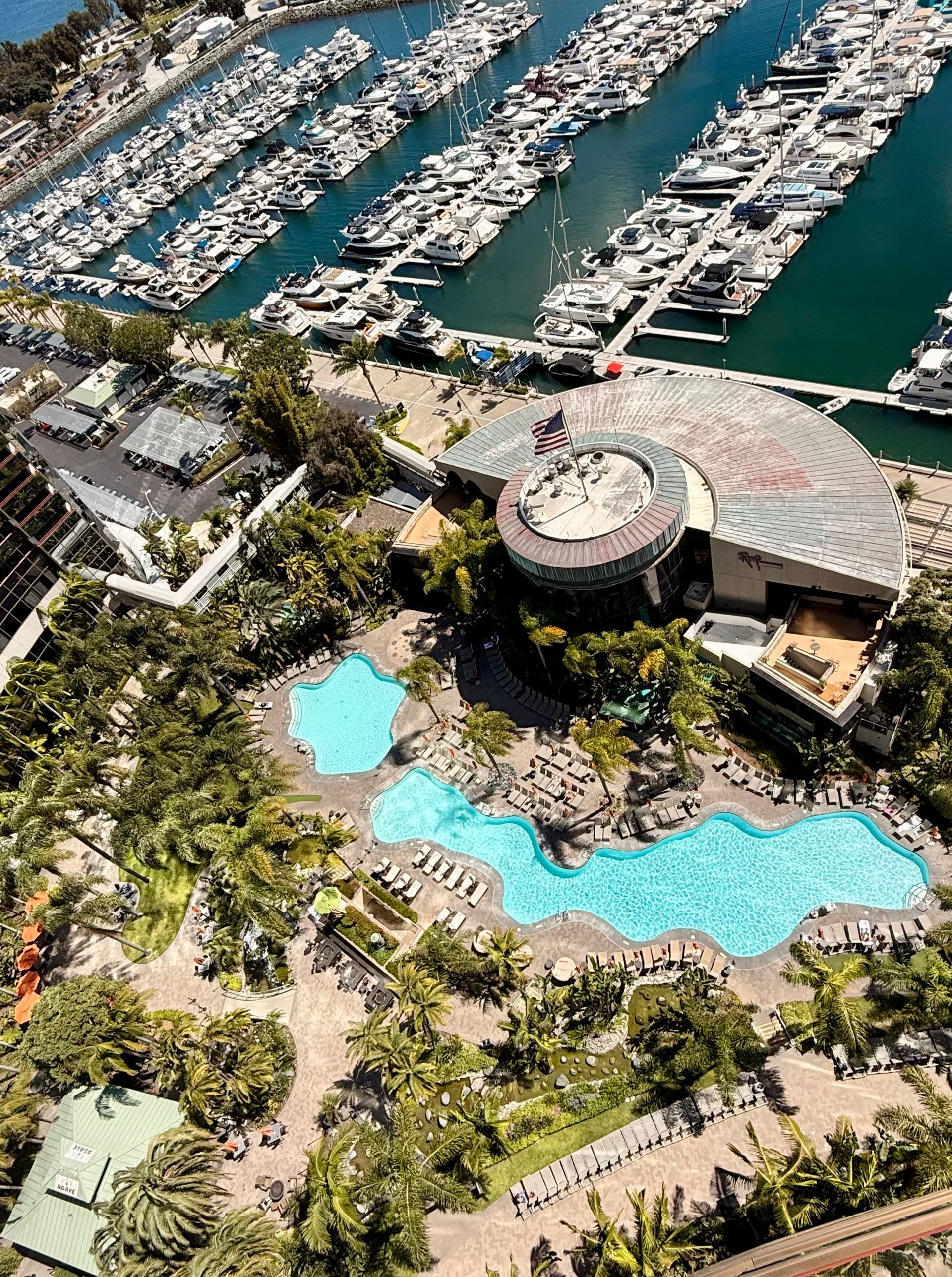 Aerial view of The Marriott San Diego Marquis marina, luxury buildings, and a turquoise resort pool surrounded by palm trees.
