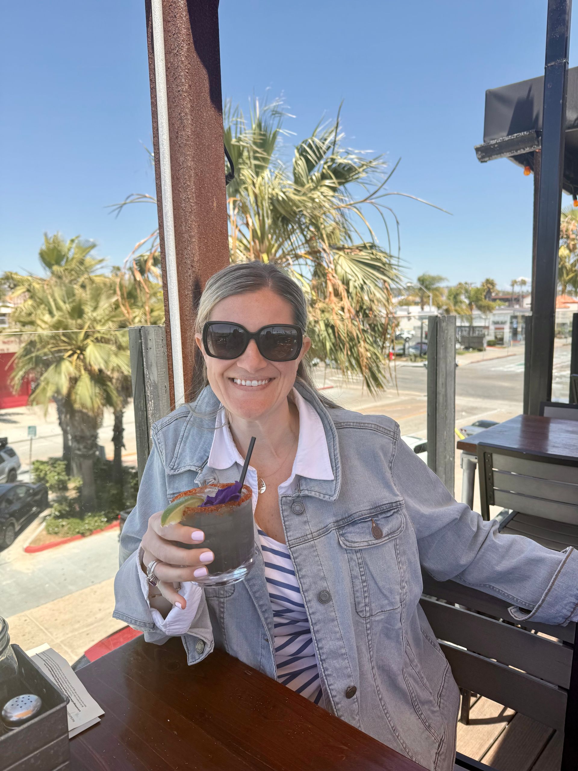 Woman in sunglasses smiling on a sunny patio, holding a drink with palm trees in the background in Pacific Beach, San Diego