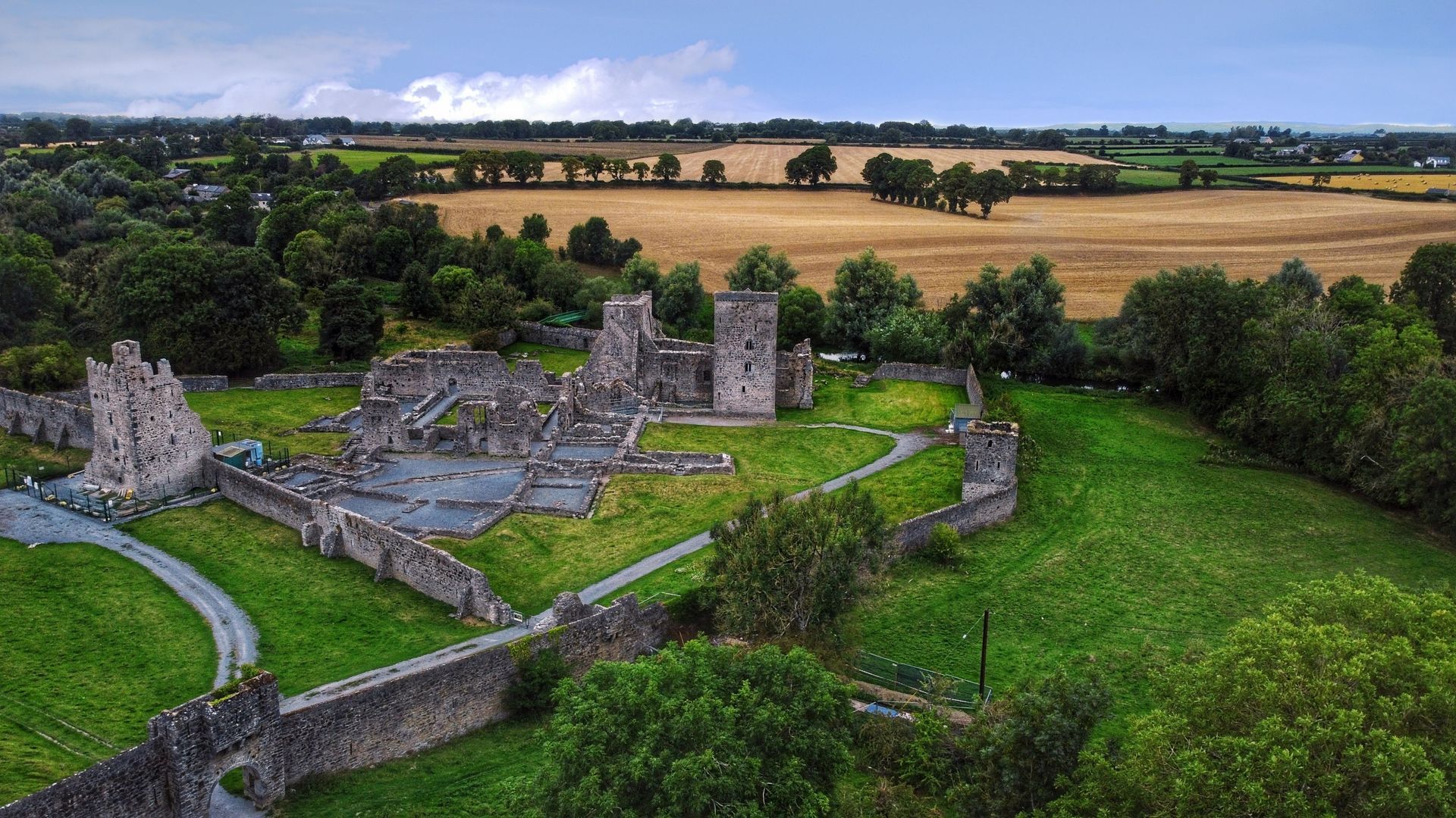 Aerial view of a ruined stone castle surrounded by green fields and trees under a cloudy sky in Ireland
