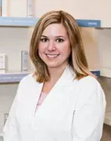 Woman in white coat, smiling, in a medical setting with shelves and products behind her.