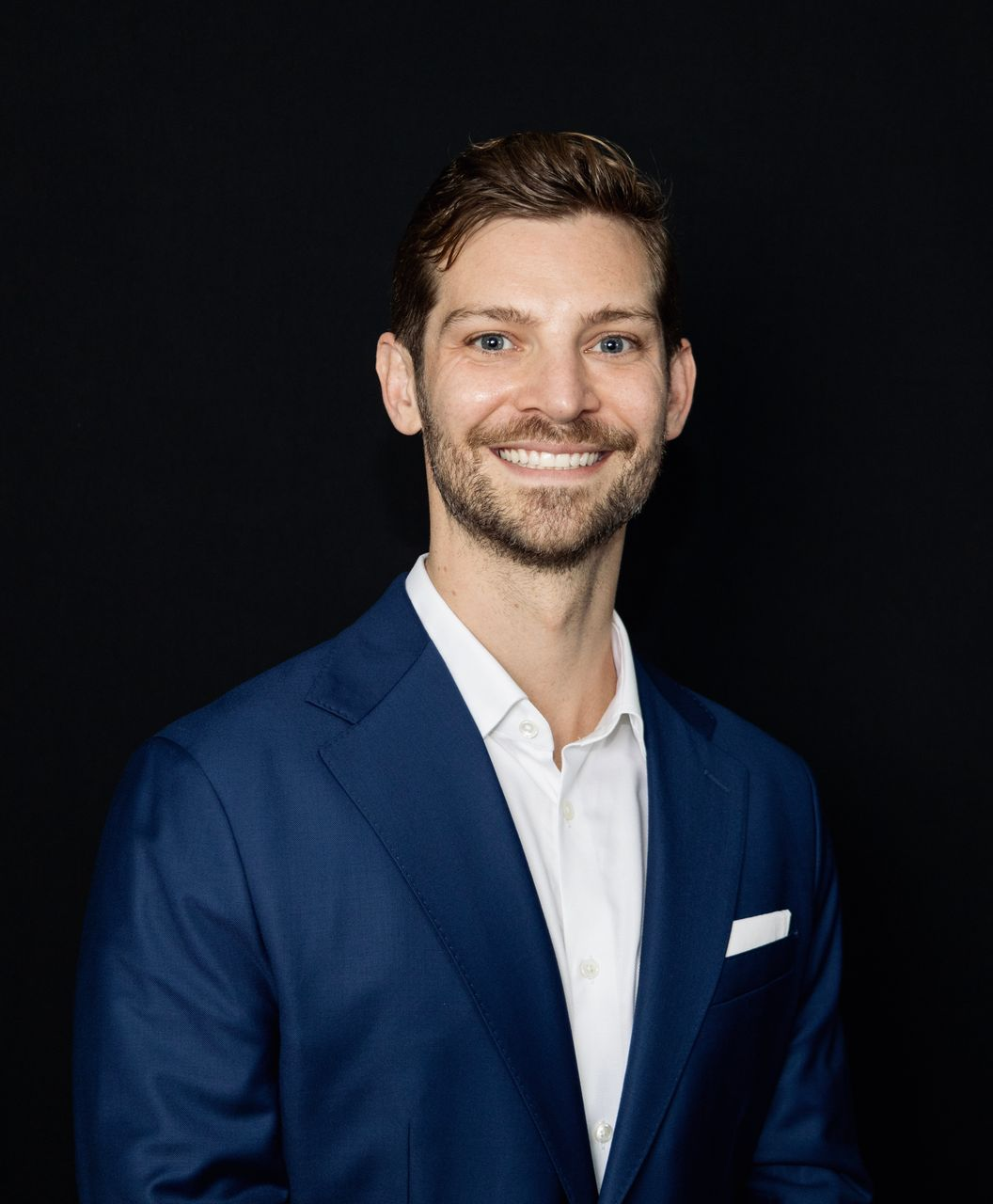 Man in blue blazer smiles, white shirt, pocket square, black backdrop.