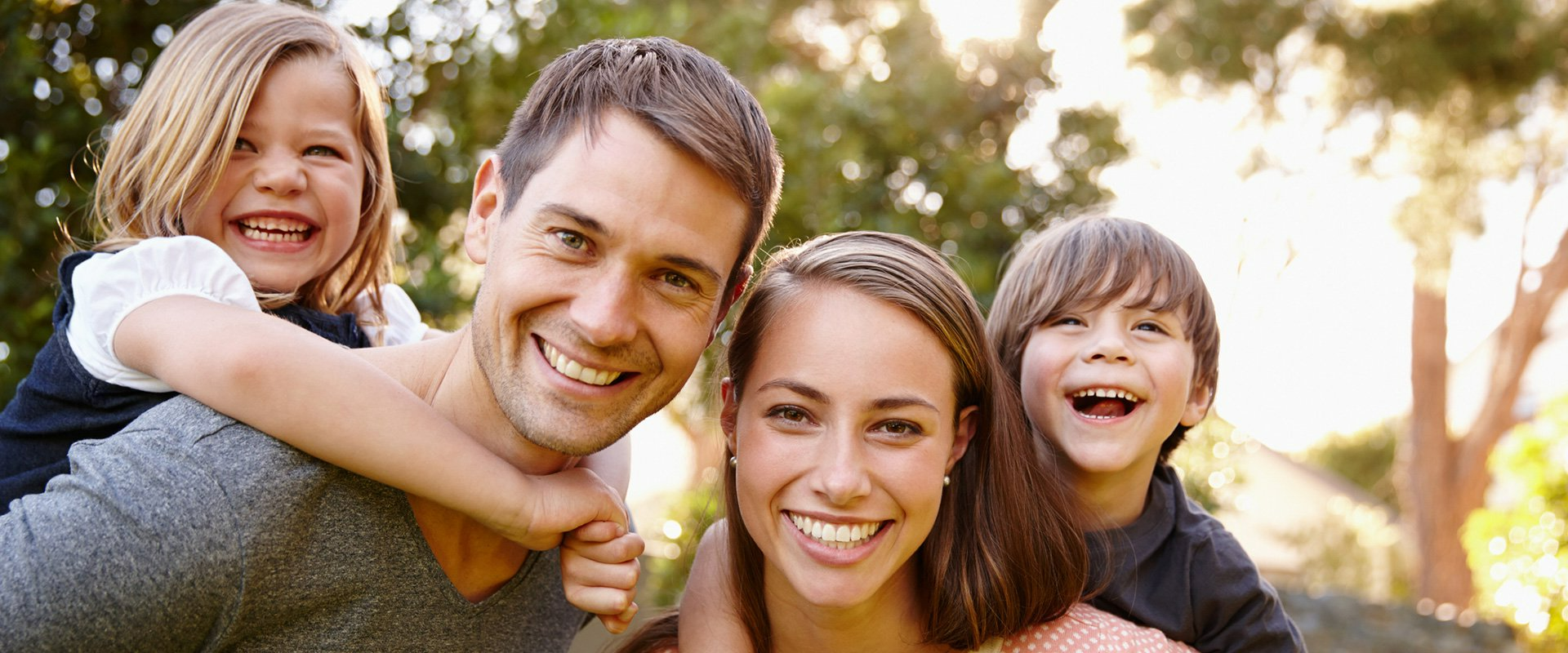 Family of four smiling widely outdoors. The dad carries one child on his back. The others smile at the camera.