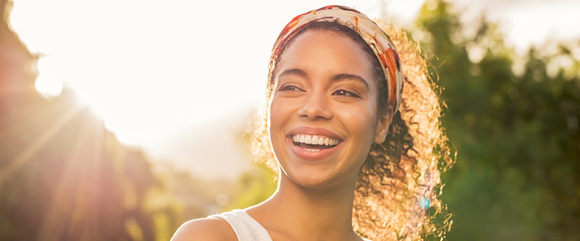 Smiling woman with curly hair wearing a patterned headscarf, outdoors in sunlight.