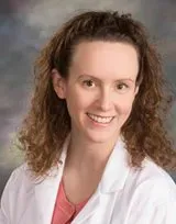 Woman with curly brown hair smiles, wearing a white lab coat over a pink shirt.