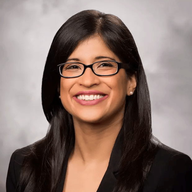 Woman with dark hair and glasses smiling at the camera, wearing a black blazer.