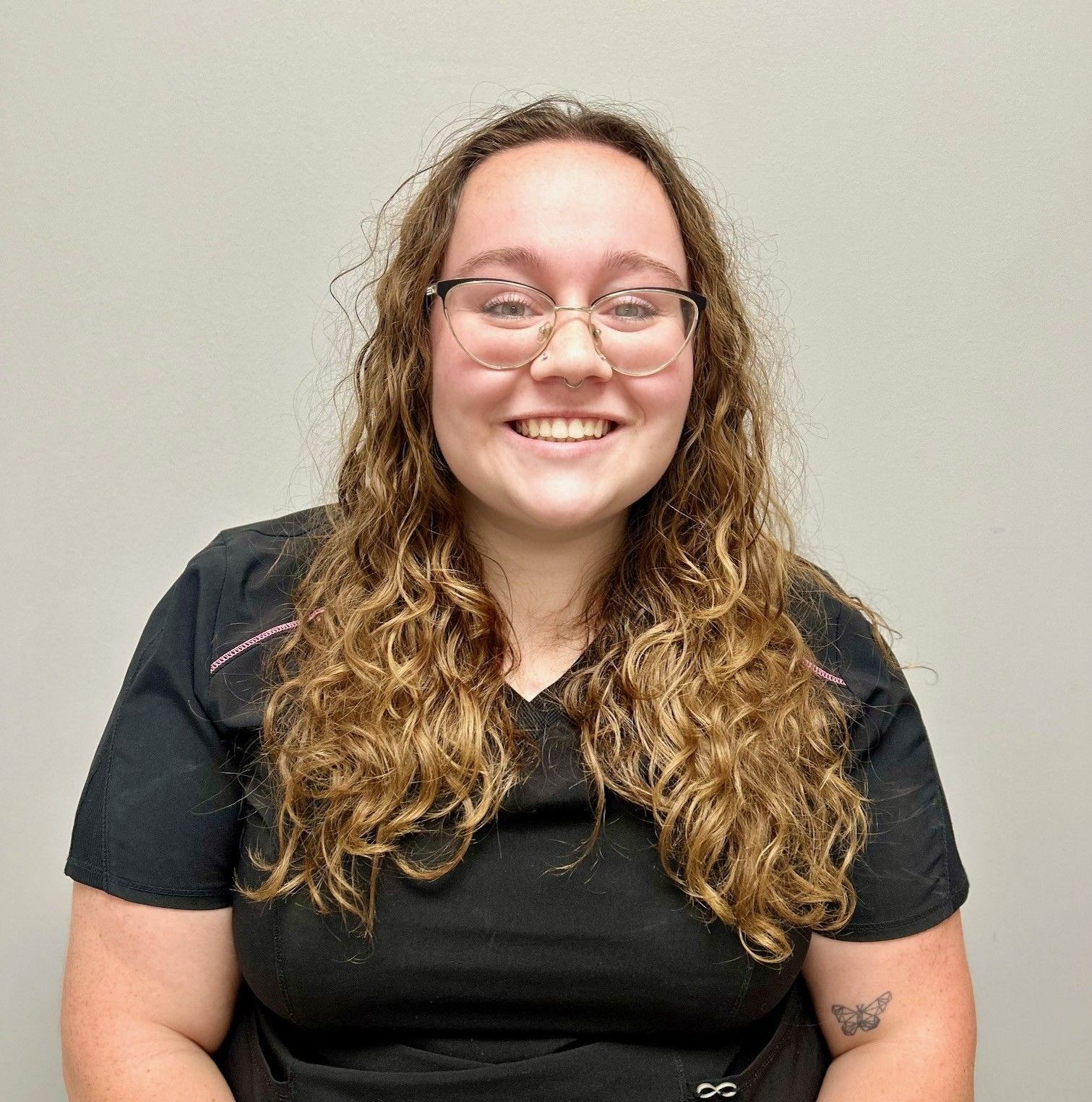 Woman with glasses smiles, long curly hair, wearing black scrubs, small tattoo.