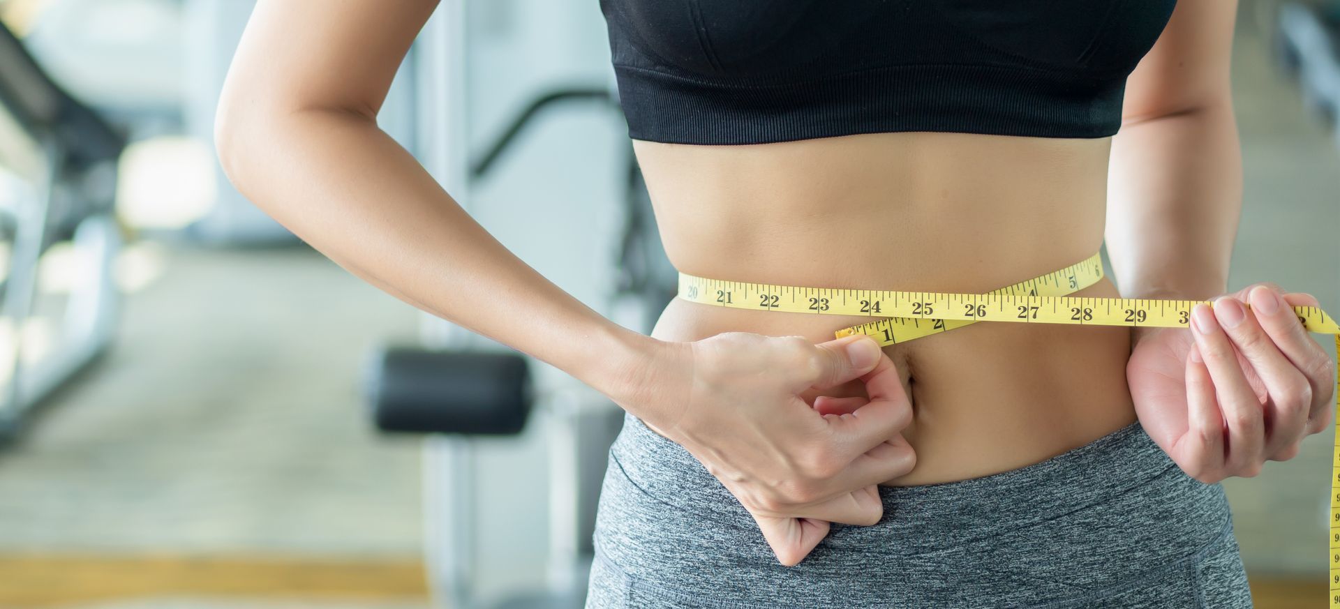 A person in workout gear using a yellow measuring tape to measure their waist in a gym setting.
