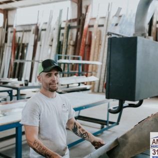 Man in a white T-shirt and black cap standing in a woodworking shop beside stacked boards and machinery