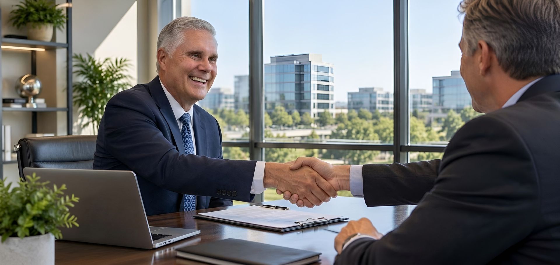 Two businessmen shake hands in a bright office with a city view.